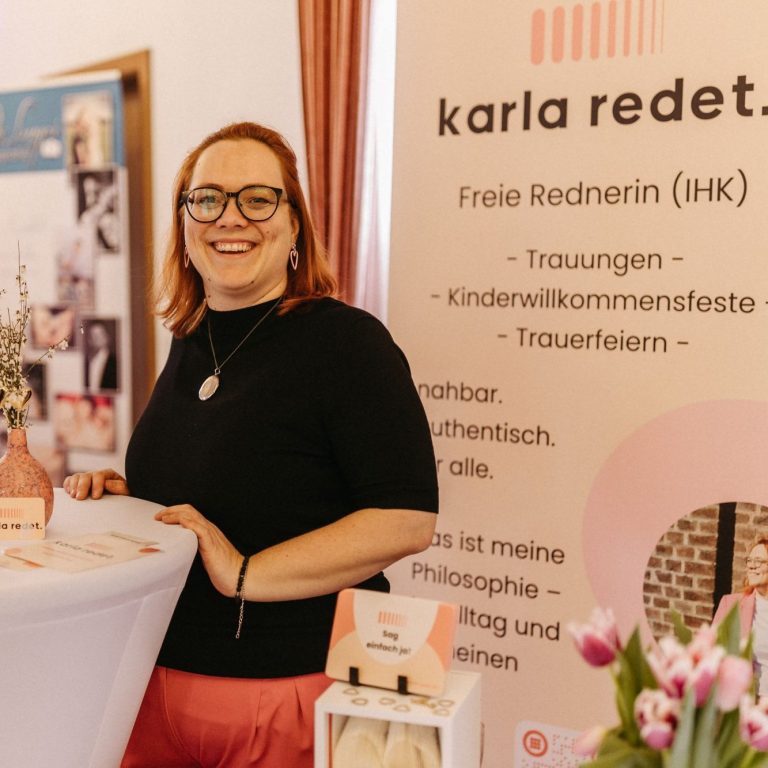 Karla bei einer Messe Karla steht an einem Messestand. Sie steht an einem weißen Tisch und wendet sich den Zuhörenden zu.